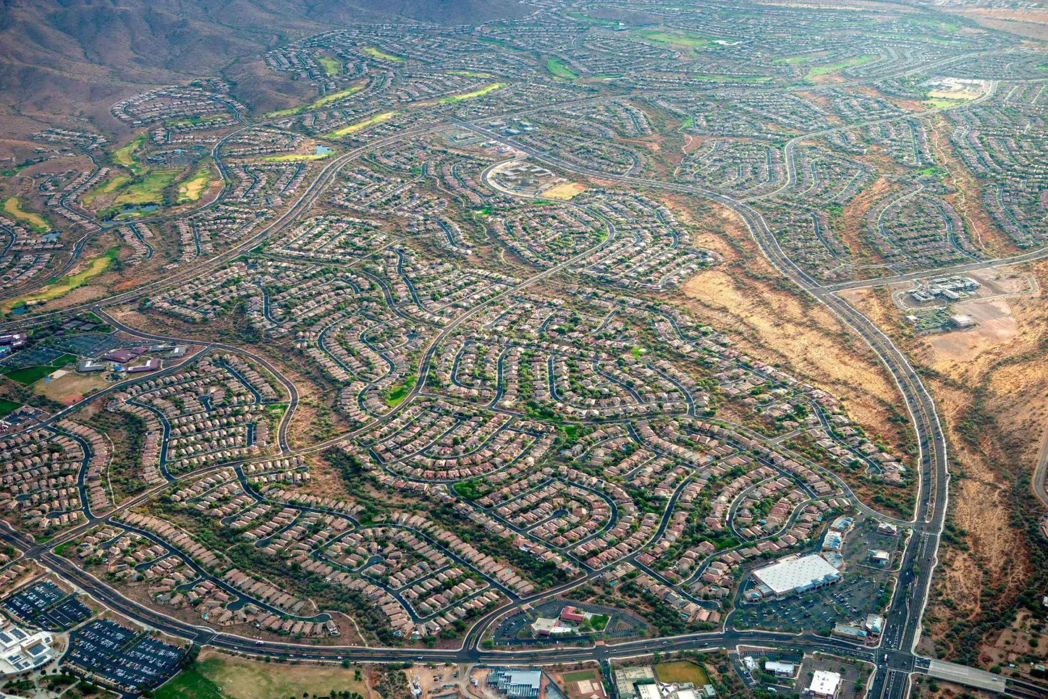 Aerial view of a sprawling suburban neighborhood with winding roads, surrounded by desert landscape and mountains, capturing urban development against natural terrain.