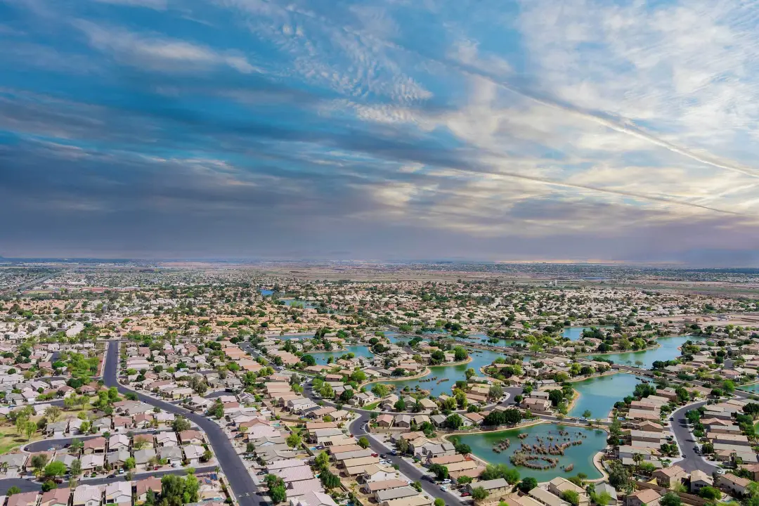 Aerial view of a suburban area with winding streets, houses, and small lakes under a vast, partly cloudy sky.
