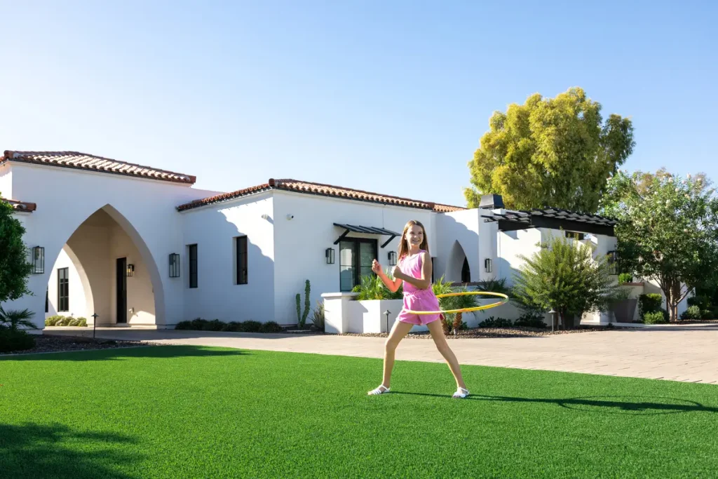 A woman in a pink top and white shorts playing tennis on a green lawn in front of a white Mediterranean-style house with arched doorways.