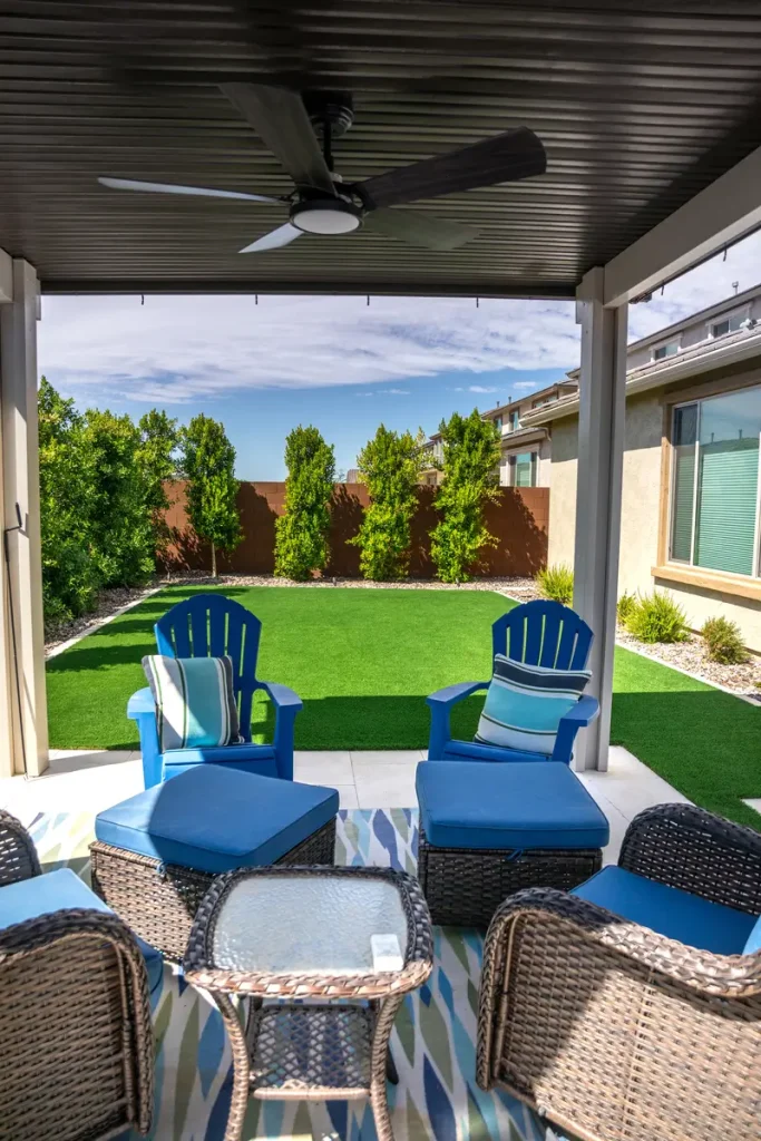 Covered patio with wicker chairs and blue cushions, a glass-top coffee table, and a manicured lawn with hedges in the background.