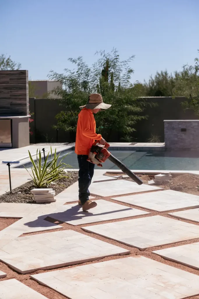 Person in an orange shirt and sunhat uses a leaf blower on a geometric stone patio beside a pool.
