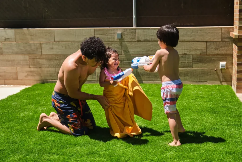 Three kids play on a sunny lawn, a boy kneels beside a girl in a yellow dress while another boy aims a water gun.